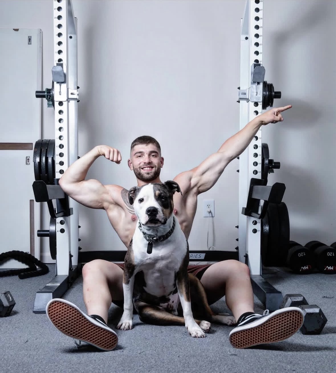 Jeffrey Heckendorn flexing in his gym with his pitbull seated in front of him