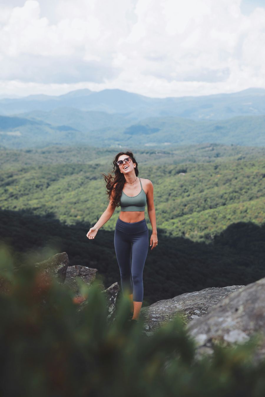 A smiling woman standing on a rocky summit overlook above forested mountains
