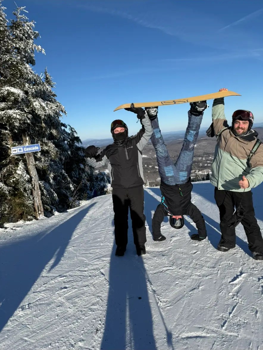 Three friends on a snowy ski run, one doing a handstand under a snowboard